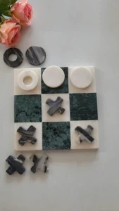 A marble tic-tac-toe board with white and dark green tiles, featuring round white pieces and dark X-shaped pieces, arranged with some pieces placed on the board and others nearby, alongside pink flowers.