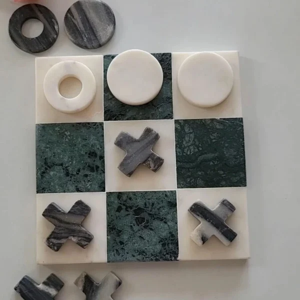 A marble tic-tac-toe board with white and dark green tiles, featuring round white pieces and dark X-shaped pieces, arranged with some pieces placed on the board and others nearby, alongside pink flowers.
